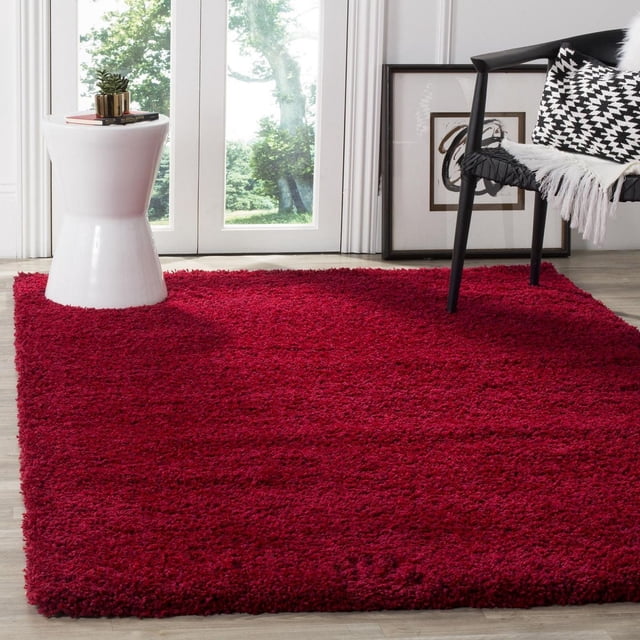 Safavieh California Solid Plush Shag Area Rug in deep red with a super dense 4.5kg/m² polypropylene pile, placed on light wood flooring in a bright room featuring a white ceramic side table with a small plant and books, and a black woven chair with a black and white geometric cushion and white throw, suitable for adding plush comfort to living areas.
