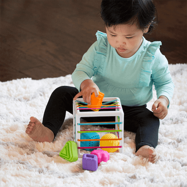 Fat Brain Toys InnyBin, un Jouet de Tri des Formes pour 10 Mois et Plus, Présentant un cube Blanc avec des Bandes Élastiques et six Formes Épaisses et Texturées aux Couleurs Vibrantes, Incluant un cube, un Diamant, une Sphère, un triangle, une Fleur et le logo Fat Brain. un Enfant Est Vu Jouant avec le Jouet sur un Tapis Blanc Doux.