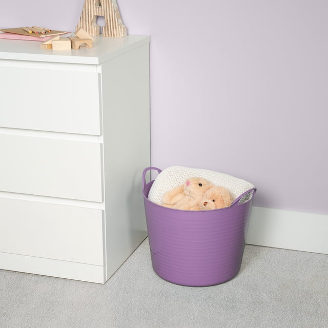 Neatfreak Kids Large Flexible Bin w/ Handles in purple soft-sided plastic with horizontal ridges, holding a white knitted blanket and two beige plush toys, placed on grey carpet beside a white three-drawer dresser against a light lavender wall, suitable for versatile home storage and organizing children's items.