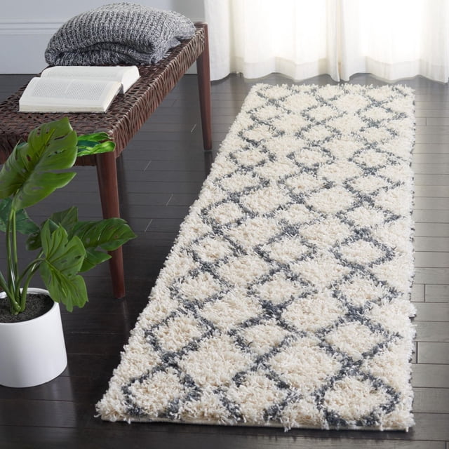 Safavieh Venus Nanda Geometric Shag Area Rug runner with a 1.77-inch medium pile, cream base and dark gray diamond pattern, placed on dark hardwood floor beside a woven bench with folded gray blankets and an open book, near a potted green plant and sheer white curtains, ideal for adding plush texture and visual interest to contemporary or boho-mod rooms.