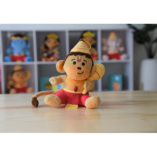 MODI TOYS Hanuman Plush (Small - 6") Mantra Singing Plush Toy featuring Baby Hanuman in a seated position with a gada in his left hand and a blessing gesture in his right. The plush has a flexible tail and a velcro strap for easy attachment to strollers, diaper bags, or car seats. It softly sings five Hanuman mantras when its belly is pressed. Displayed on a wooden surface with a blurred background of similar plush toys on shelves.