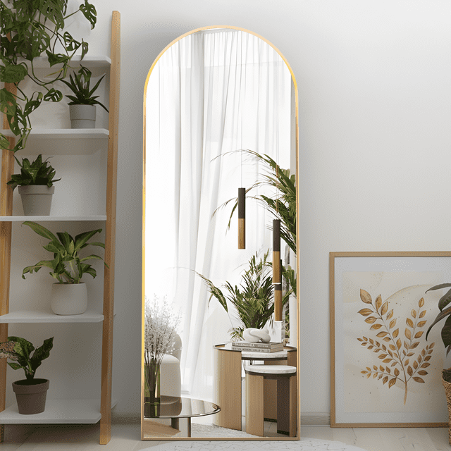 CISTEROMAN Arched Full Length Mirror measuring 64 inches by 21 inches, featuring a gold metal frame and a vertical standing design. The mirror is positioned in a modern interior setting with plants, a white curtain, and decorative elements in the background. Its arched shape and slim frame add a touch of elegance, making it suitable for bedrooms, living rooms, or corridors. The mirror includes an explosion-proof film for added safety and durability.