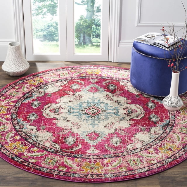 Safavieh Monaco Toria Traditional Area Rug, round with a vibrant Bohemian-chic Persian design in red, cream, blue, and yellow polypropylene pile, power-loomed for durability, displayed on a hardwood floor near glass doors with natural light, next to a blue ottoman and decorative vases, ideal for adding color and texture to living spaces.