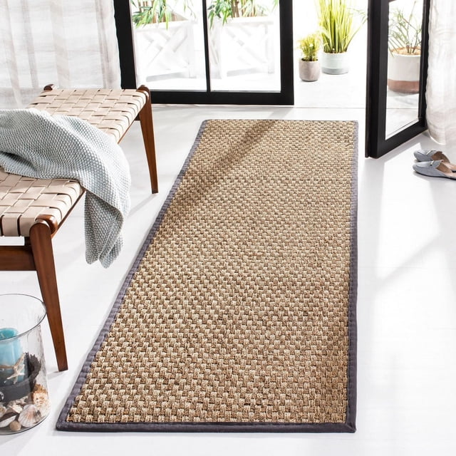 Safavieh Natural Fiber Arbor Border Area Rug featuring woven sisal texture and dark grey border, placed on a white floor in a bright room with glass doors, wooden bench with knitted throw, and potted plants, ideal for coastal or casual interiors.