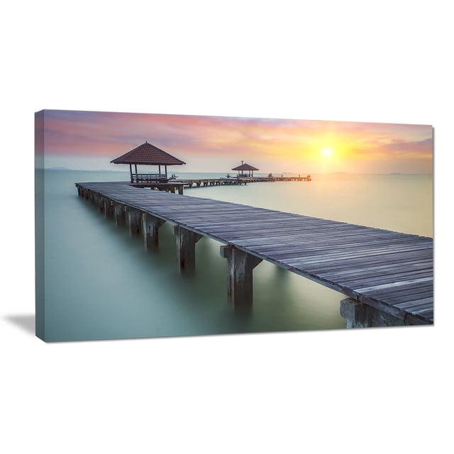 Design Art Wooden Sea Bridge And Sunset Seashore Photo Canvas Art Print featuring a gallery-wrapped canvas wall art of a wooden pier with two covered gazebos stretching over calm sea water at sunset, ideal for enhancing home décor with serene coastal imagery.