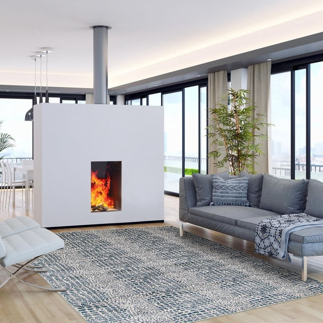 ECARPET Talia Navy Rug, an indoor accent rug with textured abstract navy and white design, placed on light hardwood flooring in a modern living room. The rug lies before a gray sofa with patterned cushions and a white chair, near a white central fireplace with visible flames and a tall green plant by large floor-to-ceiling windows.