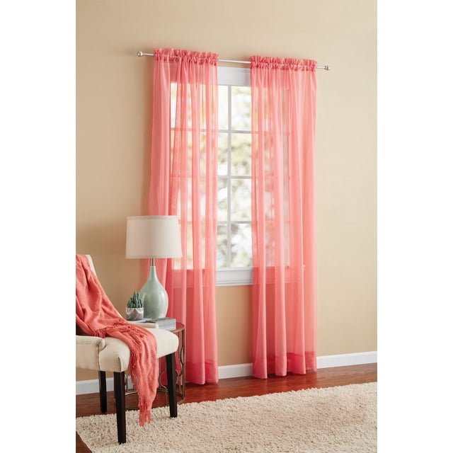 MAINSTAYS Marjorie Solid Voile Curtain Panel in coral pink sheer voile fabric hanging on a silver curtain rod over a white-framed window in a beige room with hardwood floors, next to a beige upholstered chair draped with a matching coral throw, a small round side table with a green ceramic lamp and a small cactus plant, and a cream shag area rug, ideal for light filtering and decorative window treatments and suitable for layering with heavier curtains.