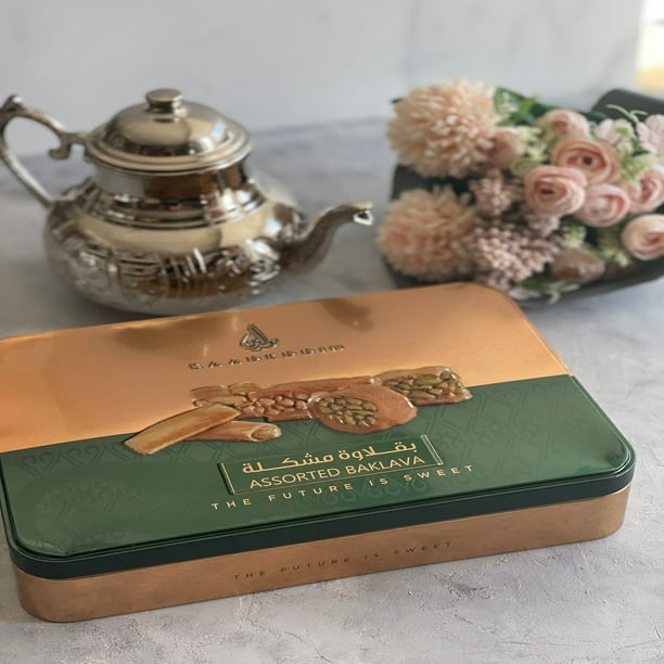 Saadeddin Baklava 500 g dans une Boîte Métallique Rectangulaire avec Couvercle de Couleur Cuivre et base Vert Foncé, Présentant des images de Noix Assorties et de Morceaux de Baklava, Placé sur une surface Gris Pâle avec une Théière Argentée et un bouquet de Fleurs Roses, Convient aux Réunions de Famille Ou aux Fêtes.