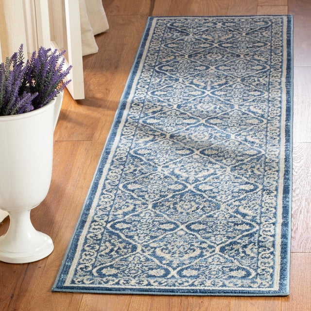 SAFAVIEH Brentwood Naomh Geometric Area Rug with blue and cream geometric patterns and a decorative border, crafted from soft synthetic yarns in a close cut pile, shown on a hardwood floor next to a white vase with purple flowers, suitable for enhancing traditional or transitional home decor.