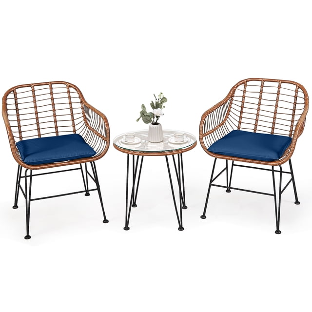Patiojoy 3PCS Patio Rattan Furniture Set featuring a round tempered glass top table and two rattan armchairs with navy cushions. The set includes a brown PE wicker design with black steel frames, ergonomic curved backrests, and non-slip feet for stability. The table is adorned with a vase and cups, showcasing its use for outdoor settings like patios, gardens, or poolside areas.