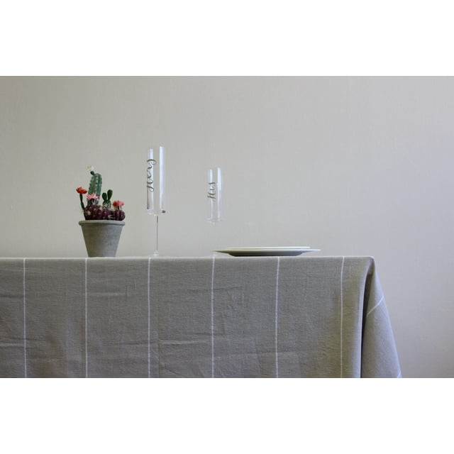 Fabstyles Fouta 100% Cotton Stripe Table Cloth in grey with thin white vertical stripes on a rectangular table, styled with a small potted cactus, two clear champagne flutes with gold lettering, and stacked white plates, ideal for indoor dining and adding modern colour to table linens.
