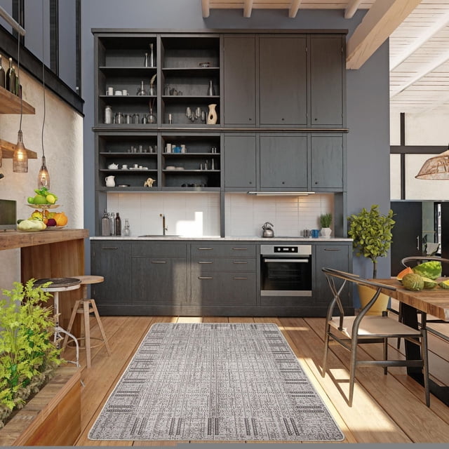 ECARPET Kitchen, Hallway, Laundry Room, Entrance, Kids Room, Bathroom Mat & Utility Rug in grey with geometric bordered pattern, made of durable polypropylene with non-slip rubber backing, placed on light hardwood floor in a modern kitchen featuring dark grey cabinets, open shelving with kitchenware, stainless steel oven, and natural sunlight.