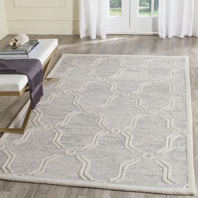 SAFAVIEH Cambridge Leland Faded Geometric Wool Area Rug with hand-tufted wool texture in faded grey and raised cream geometric patterns. Displayed on light wood flooring near a white bench with gold legs and dark purple throw, suitable for modern home or office decor and as a soft accent in seating areas.