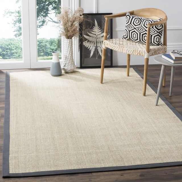 Safavieh Natural Fiber Forrester Border Area Rug with natural woven sisal texture and dark grey border on dark hardwood floor, styled with a wooden chair with woven seat and geometric cushion, small grey side table with books and glasses, and decorative vases with dried grasses near a window. Often used in coastal or casual beach house style interiors.