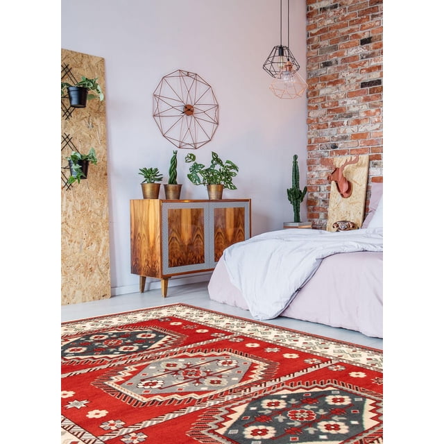 ECARPETGALLERY 100% Wool Traditional Indoor Area Rug with bold geometric Persian Kazak-inspired patterns in red, grey, and cream tones, displayed in a bedroom with a wooden cabinet holding potted plants, a white bed with light bedding, exposed brick wall, and modern wireframe pendant lights, also suitable for entrance, living, or dining rooms.