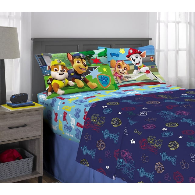 Paw Patrol Boy "So Fun" Full Sheet Set on a bed with gray headboard in a bright room. The set includes a fitted sheet and two pillowcases featuring Paw Patrol characters Marshall, Rubble, Chase, and Skye on colorful backgrounds with stars, paw prints, and badges. The flat sheet and comforter have a blue pattern with multicolored Paw Patrol icons, bones, and paw prints. Made of 100% polyester and machine washable, this bedding adds a playful theme to a child's bedroom.