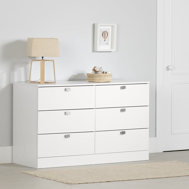 South Shore Spark Commode à 6 Tiroirs pour Enfant en Blanc Pur avec six Tiroirs Spacieux sur Chemins en Métal et Poignées Carrées en Métal, Montrée Orientée vers l'Avant contre un Mur Gris Pâle. la Commode Présente une Lampe de table beige et un Panier Tressé avec des Peluches sur le Dessus, Placée sur un Plancher Clair avec un Tapis beige, Convient pour Organiser les Vêtements d'Enfants et les Articles de Chambre.