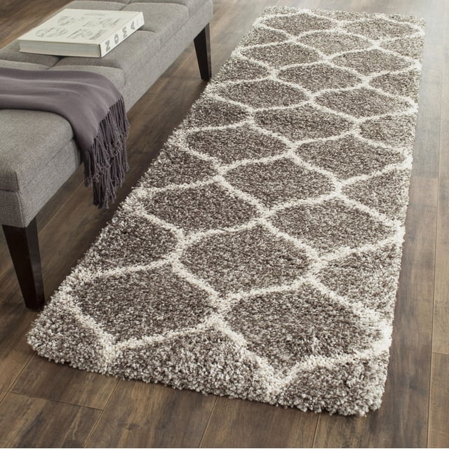 Safavieh Hudson Tapis Shag Géométrique Arline Exposé sur Plancher de Bois Franc à Côté d'Un Banc Rembourré Gris avec un Jeté Violet et un Livre. le Tapis Présente une Épaisse, Pelucheuse pile Shag avec des Motifs de Carreaux taupe et Ivoire en Forme d'Oignon, Convient pour Ajouter de la texture à un décor Urbain Ou Moderne et durable pour les Zones à Fort Passage.