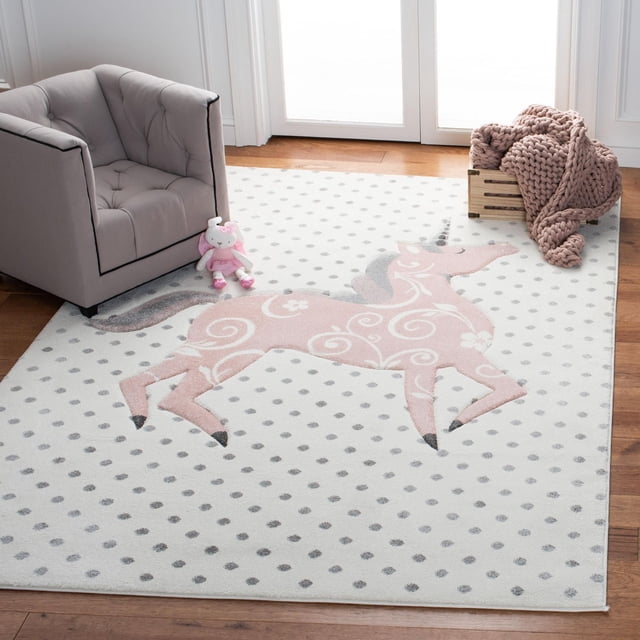Safavieh Carousel Kids Dalach Unicorn Area Rug with a front-facing view of a durable synthetic yarn kids rug featuring a large pink unicorn with white floral swirls on a white background with gray polka dots, placed on a hardwood floor beside a gray tufted kids armchair, a pink stuffed animal, and a wooden crate holding a chunky pink knit blanket, ideal for adding soft, playful decor to children's rooms.