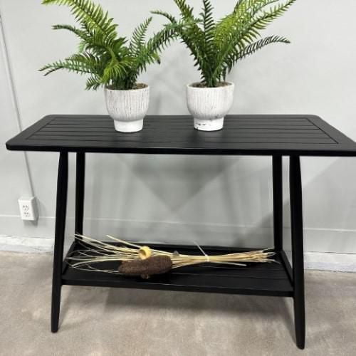 Windsor console table 15''x39' made of black acacia wood with a slatted top and lower shelf, shown with two white ceramic pots holding green ferns on top and dried decorative grasses on the shelf below, suitable for outdoor patio or indoor use as a sideboard or accent table.