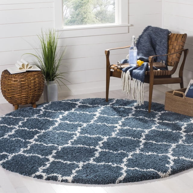Safavieh Hudson Jaye Geometric Shag Area Rug, round with a thick plush shag pile in blue and ivory geometric tile pattern, placed on light hardwood floor in a modern room with a wooden armchair draped with a blue throw, a wicker side table with a book, and a green plant, suitable for adding texture to living areas.