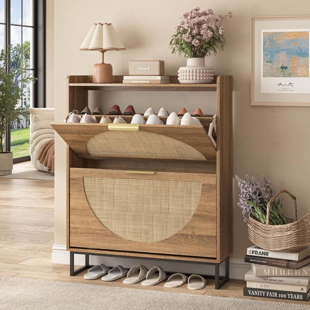 Vabches Rattan Shoe Storage Cabinet with 2 Flip Drawers, featuring a freestanding narrow design with metal legs. The cabinet showcases a natural wood finish with hand-woven rattan panels and a geometric half-round design on the flip doors. It is displayed in an entryway setting, holding multiple pairs of shoes in its spacious compartments, with additional slippers stored underneath. The top surface is styled with decorative items, including a lamp, books, and a vase with flowers. The cabinet is crafted from engineered wood and metal, offering a space-saving solution for entryways or hallways.