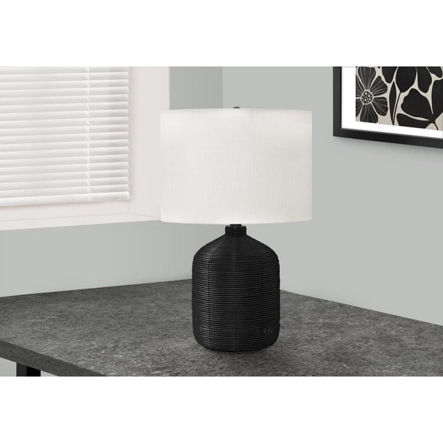 Monarch Specialties Lighting 23-inch modern table lamp with a black rattan ginger jar-shaped base and an ivory linen drum shade, featuring a 3-way switch, placed on a dark grey stone tabletop near a window with white blinds and a black and white floral framed print on a light grey wall.