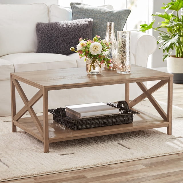 Mainstays Farmhouse Coffee Table with rustic weathered oak finish and X-brace design on each end, featuring a spacious top surface holding decorative glass vases and a flower arrangement, plus a bottom shelf with a woven basket and books, set on a cream textured rug in a bright living room with a white sofa and cushions. Its sturdy design is also suitable for use as a versatile workspace or display surface in a den or office.