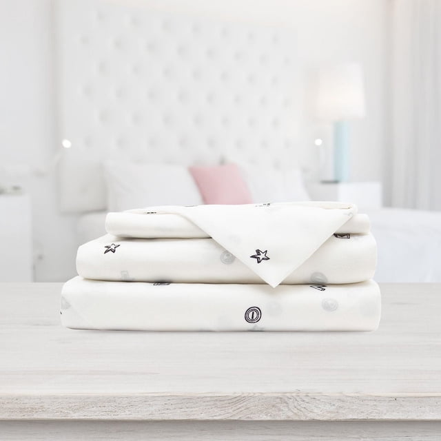 Safdie & Co. 3-piece twin size microfiber sheet set featuring white fabric with small black gaming-themed icons, displayed folded on a light wood table in a bright bedroom.