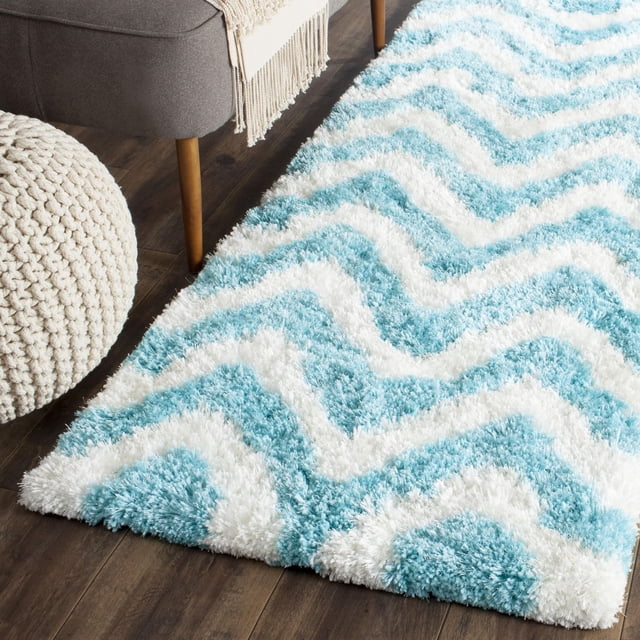 Safavieh Barcelona Carmella Zigzag Stripes Shag Area Rug with plush light blue and white zigzag stripes on a dark hardwood floor, shown beside a knitted pouf and mid-century modern chair with wooden legs and beige throw, suitable for adding soft texture and Mediterranean style to runner rug areas.