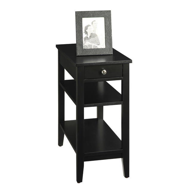 Convenience Concepts American Heritage Three Tier End Table with Drawer in black. Features a sleek black wood finish, a single drawer with a round silver knob for hidden storage, and two open shelves for additional storage. Made from MDF, birch veneer, and mixture veneer. Dimensions: 23.5 inches wide, 11.25 inches long, and 24 inches high. A framed photo is displayed on the tabletop.