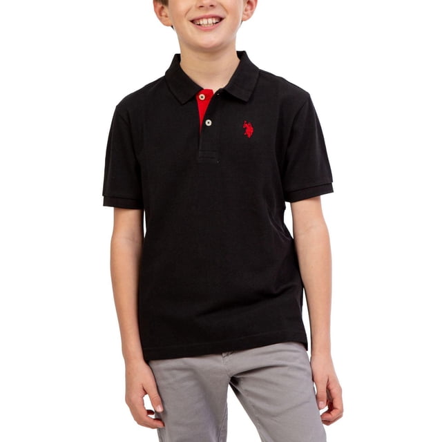 U.S. Polo Assn. Boy's Short Sleeve Polo Shirt in black with a red inner placket and embroidered red logo on the left chest, featuring a fold-over collar, short sleeves, and side vents at the hem, paired with light grey pants, suitable for school or casual wear.