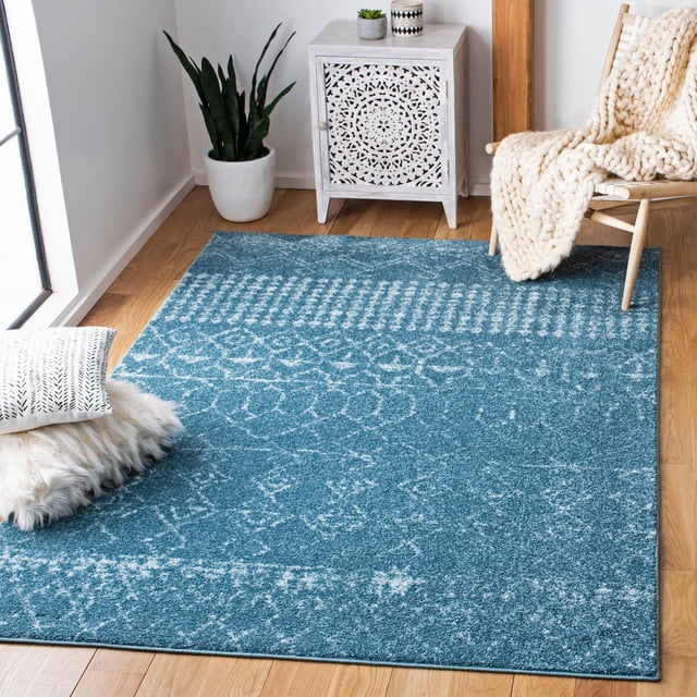 Safavieh Tulum Coriander Moroccan Geometric Area Rug in blue with white geometric patterns, hand-knotted wool with plush texture, placed on light hardwood floor in a bright room with a white carved wooden cabinet, a snake plant in a white pot, a wooden chair draped with a chunky cream knit blanket, and a fluffy white floor cushion with a patterned pillow, ideal for adding warmth and style to contemporary home décor.