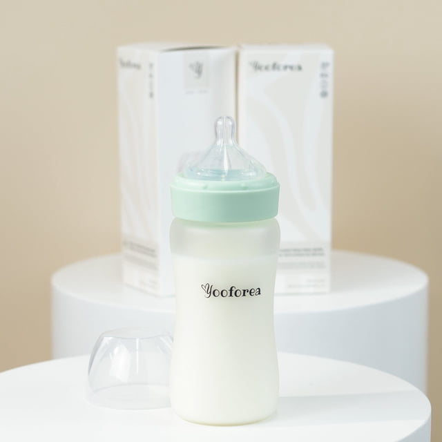 Yooforea Silicone Coated Glass Baby Bottle, 6M+ Fast Flow Nipple, shown in a 270 ml (9 oz) size with a tea-colored medical-grade silicone coating for shatter protection. The bottle features a wide neck for easy cleaning, a stable base, and a soft, anti-colic nipple designed to mimic natural breastfeeding. The image highlights the bottle with a mint green collar and transparent nipple, placed on a white surface with Yooforea packaging in the background.