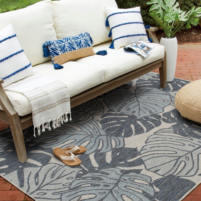 Hayweld Blue and Grey Woven Area Rug with navy and steel grey tropical leaf pattern on dusky blue and ivory background, placed on a brick patio beneath a wooden outdoor sofa with cream cushions and blue tassel pillows, featuring a woven throw and sandals, suitable for indoor or outdoor spaces.