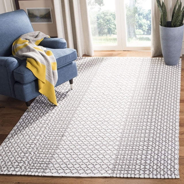 Safavieh Montauk Cara Tapis Géométrique sur Plancher de Bois dans un Salon Lumineux, avec texture en Coton Tissé à Plat et Motif Géométrique Gris et Blanc. Positionné sous un Fauteuil Bleu avec un Jeté à Rayures Grises et Jaunes, Près de Grandes Fenêtres avec des Rideaux Beiges et un Grand Cache-Pot Gris. Utilisé comme Tapis Coureur dans les Intérieurs Côtiers Ou Contemporains.