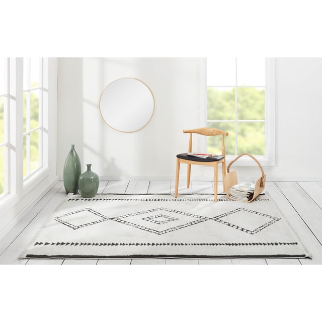 ECARPET Zelda Contemporary Area Rug for Living Room, Bedroom, Dining Room, Entrance, Hallway and More with a white background and black southwestern-inspired geometric diamond and triangle patterns, placed on whitewashed wood flooring in a sunlit room featuring a wooden chair with black cushion holding magazines, two green ceramic vases, and a woven basket with magazines, suitable as an accent rug or decorative floor covering.