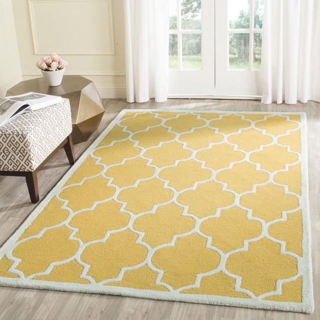 Safavieh Cambridge Kevin Geometric Area Rug with yellow hand-tufted wool and white quatrefoil pattern, displayed on light wood flooring near a patterned bench and gold geometric side table in a bright room with white curtains and glass doors, ideal for modern home decor.