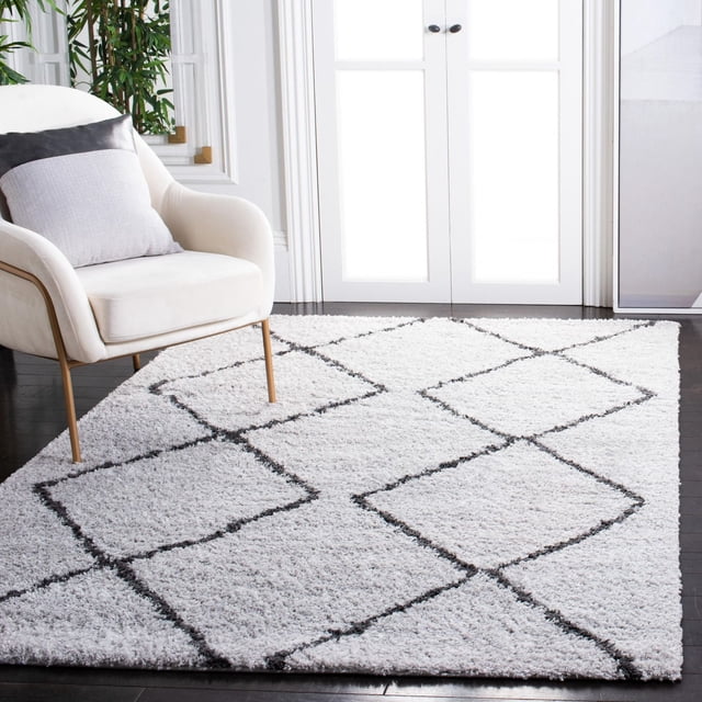 SAFAVIEH August Melanie Geometric Shag Area Rug featuring a plush white polypropylene shag pile with black geometric diamond pattern, displayed on dark hardwood flooring beside a cream armchair with a two-tone cushion, ideal for adding softness and modern style to home interiors.
