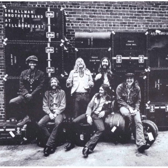 The Allman Brothers Band - At Fillmore East (vinyl) album cover featuring six band members posed casually in front of a brick wall with stacked black road cases, one displaying the album title in white text, evoking a classic live music setting.
