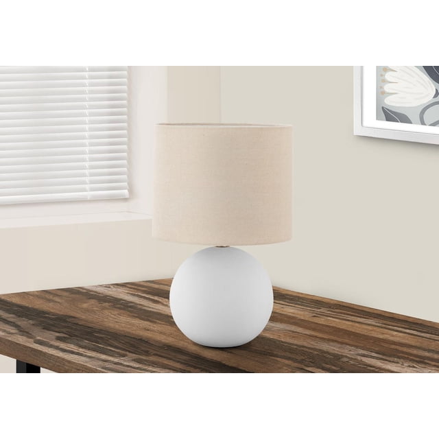 Monarch Specialties Lighting 16"h table lamp with a white ceramic globe base and cream fabric drum shade on a rustic wooden table near a window with white blinds and framed floral wall art, suitable for living rooms or bedrooms.