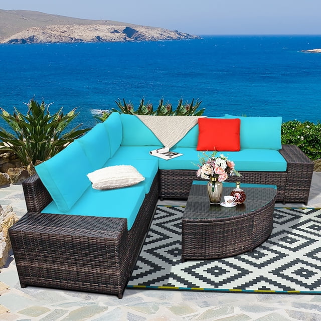Costway 6PCS Rattan Furniture Set Conversation Cushioned Sofa Armrest Garden Turquoise displayed in an outdoor setting overlooking the ocean. The set includes a sectional sofa with turquoise cushions, a corner chair, two single arm chairs, two armless chairs, and a curved glass-top coffee table. The furniture is made of all-weather PE rattan with a powder-coated steel frame for durability. The thick cushions feature removable, washable covers. The setup is placed on a patterned outdoor rug, surrounded by greenery and a scenic ocean view.
