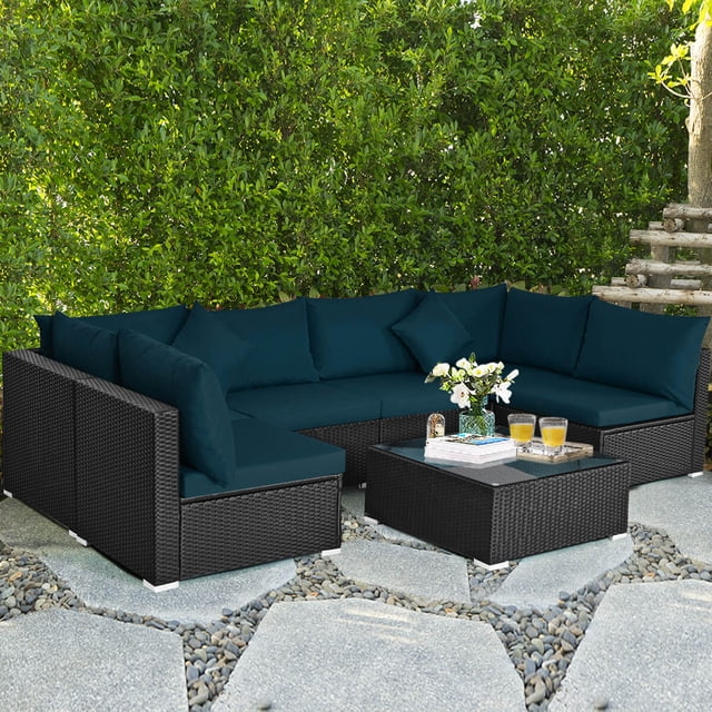 Costway 7PCS Patio Rattan Sofa Set in peacock blue, including four armless sofas, two corner sofas, and a coffee table with a tempered glass top. This outdoor sectional furniture set features a modern PE wicker frame, thick cushions, and is ideal for patios, gardens, or poolside areas. Arranged on a stone patio surrounded by greenery.