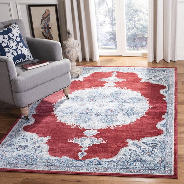 SAFAVIEH Brentwood Harland Floral Bordered Area Rug with a red central medallion and blue floral geometric patterns, made of soft synthetic yarns in a close cut pile, placed on hardwood flooring beside a grey armchair with a blue and white cushion in a bright room with large windows and beige curtains, ideal for traditional and transitional decor.