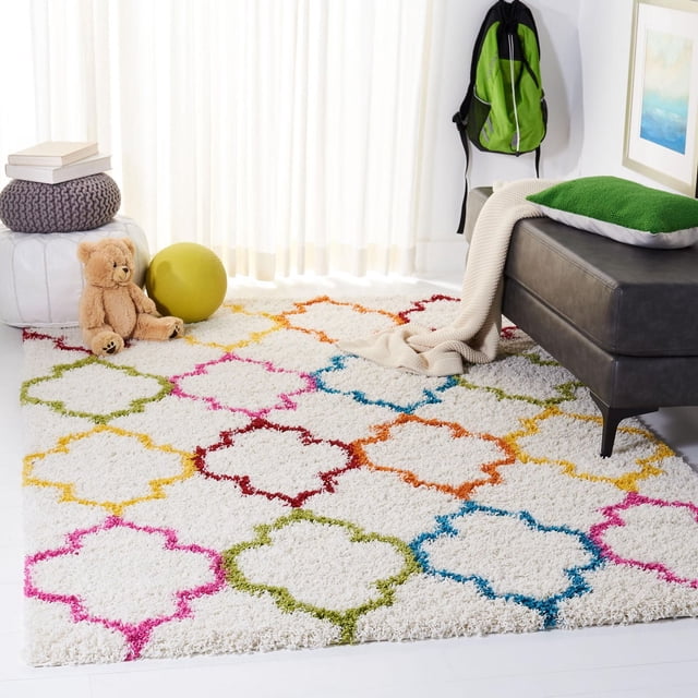 Safavieh Shag Kids Yadira Geometric Quatrefoil Area Rug with a white wool shag pile and colorful quatrefoil patterns in pink, green, blue, orange, and red, displayed in a children's room with a teddy bear, yellow ball, knitted pouf, and a dark gray bench with a green pillow and beige throw, suitable for brightening kids' bedrooms or playrooms.