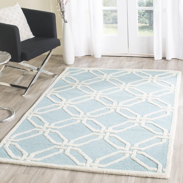 Safavieh Cambridge Kierra Geometric Area Rug displayed on a light wood floor, featuring a light blue hand-tufted wool surface with a raised white geometric chain-link pattern, set in a bright room with white curtains and a black chair with chrome legs, ideal for adding modern American style and texture to home or office decor.