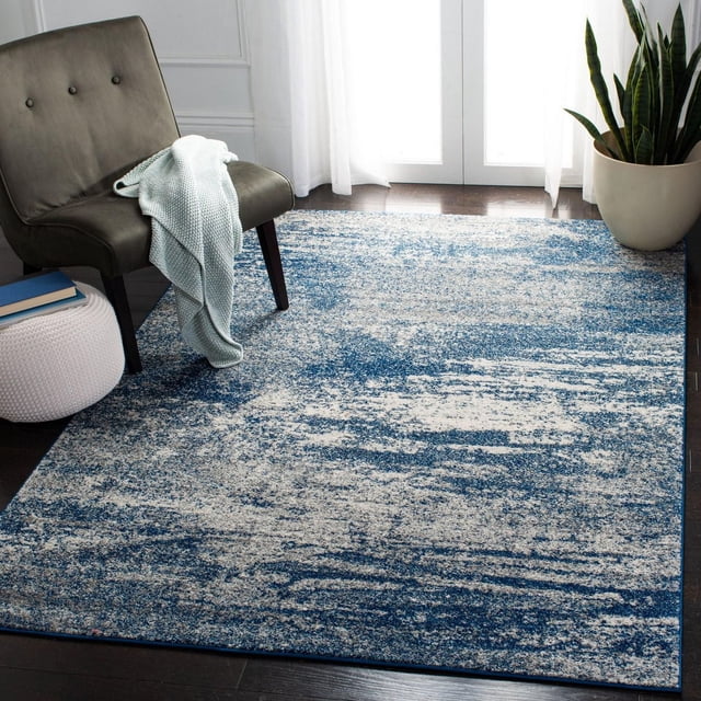 Safavieh Evoke Frosina Abstract Area Rug in navy and ivory with a power-loomed polypropylene frieze texture and modern abstract pattern, placed on dark hardwood floor beside a gray upholstered chair with a light blue knitted throw, near a white pouf with books and a tall snake plant by sheer white curtains, ideal for living room or bedroom accent.