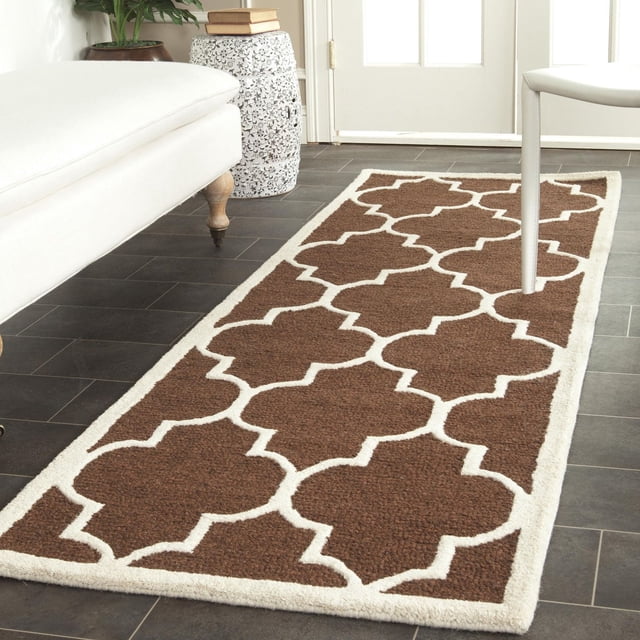 Safavieh Cambridge Kevin Geometric Area Rug in brown wool with a white geometric trellis pattern and border, displayed on dark tile flooring between a white bench and chair near a door, ideal for adding durable classic style to home or office interiors.