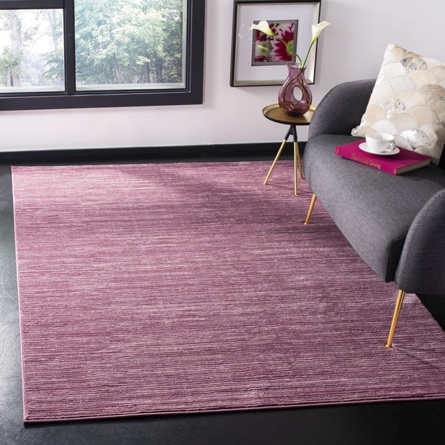 Safavieh Vision Adrasteia Overdyed Solid Area Rug in muted mauve with subtle horizontal texture, placed on a dark floor in a contemporary living room featuring a gray accent chair with gold legs, a round side table with a purple vase holding white calla lilies, and framed floral artwork. This indoor accent rug adds warmth and texture to modern living spaces.