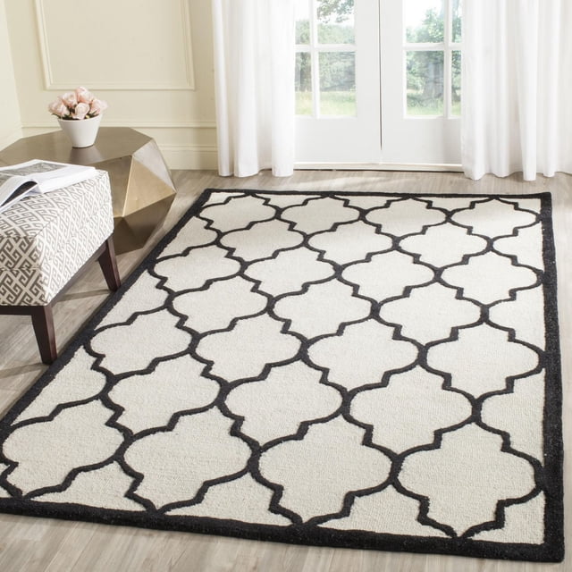 Safavieh Cambridge Kevin Geometric Area Rug in a bright room with light wood flooring, featuring a white hand-tufted wool surface with a black geometric trellis pattern and black border, styled with a patterned bench and a gold geometric side table with a white vase of pink roses, ideal for adding modern American style to home or office decor.